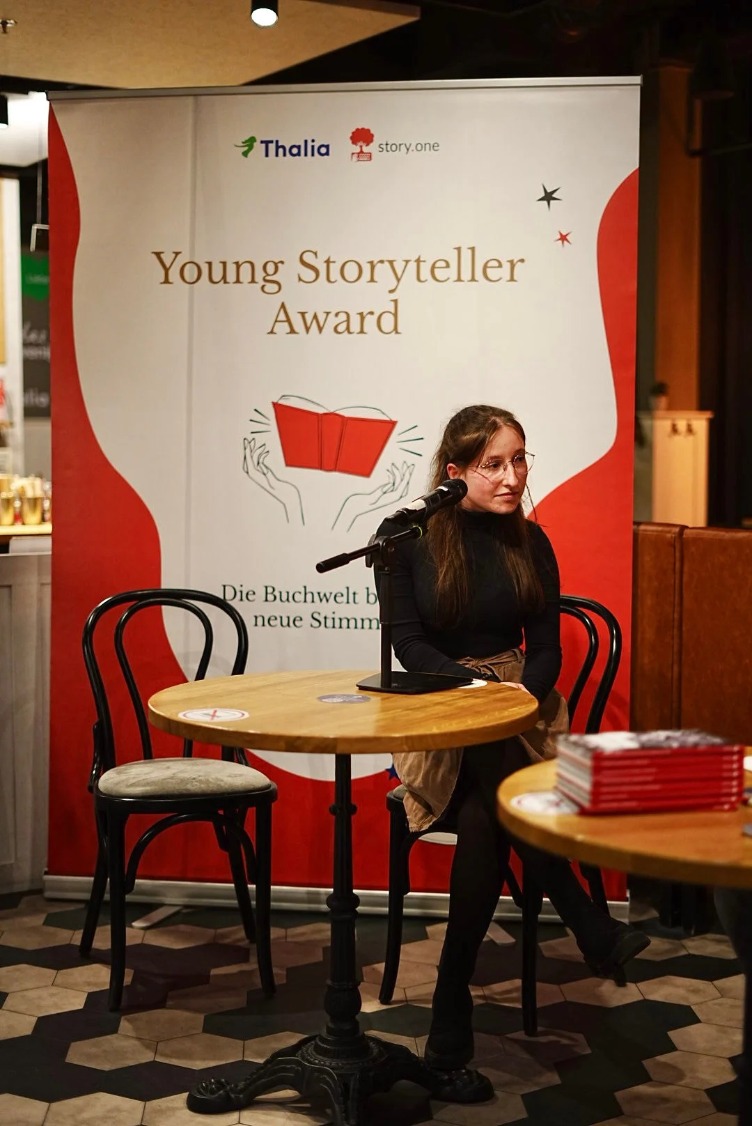 Eine Autorin sitzt am Mikrofon vor dem Banner des Young Storyteller Awards.