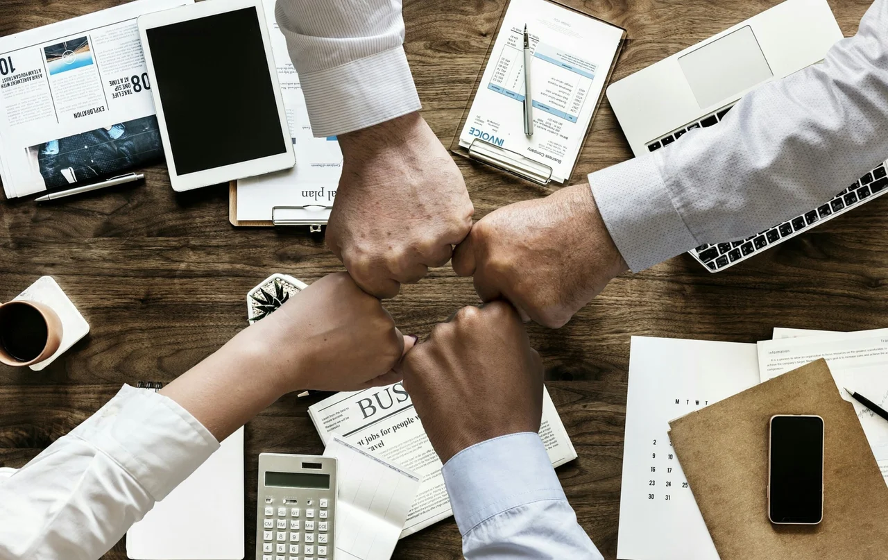 Vier Menschen führen ihre Fäuste über einem Arbeitstisch als Zeichen der Zusammenarbeit zusammen.